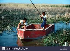 two boys fishing in a red boat on the water with tall grass and reeds behind them