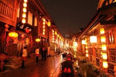 people are walking down the street at night with lanterns hanging from buildings on both sides