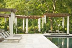 an outdoor swimming pool surrounded by white pillars and wooden pergols with chairs around it