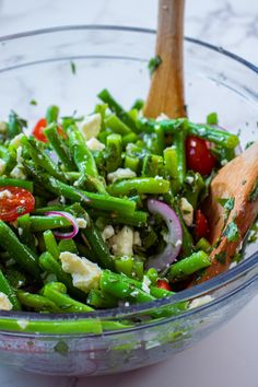 a glass bowl filled with green beans, tomatoes and other veggies next to a wooden spoon