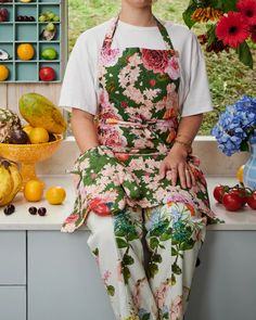 a woman wearing an apron sitting on a counter with fruits and flowers in the background