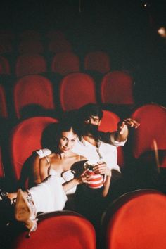 two people sitting in the middle of an empty auditorium with red seats and one person holding a drink