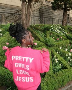 a woman wearing a pink sweatshirt standing in front of some bushes with flowers on it