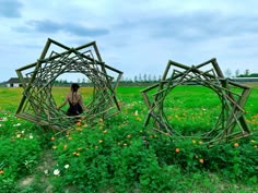 a woman standing in the middle of an open field with two wooden structures made out of sticks