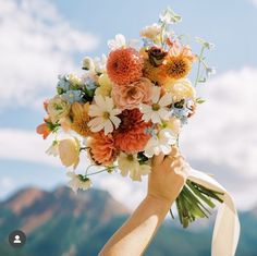 a person holding a bouquet of flowers in front of a mountain range with blue sky and clouds