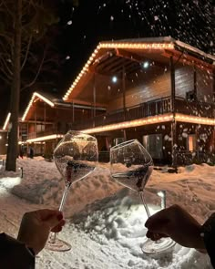 two people toasting wine glasses in front of a building at night with snow falling on the ground