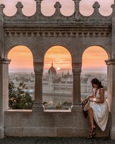 a woman sitting on top of a stone wall next to an arch in front of a city