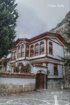 an old white and brown house with red trim on the windows, in front of a mountain