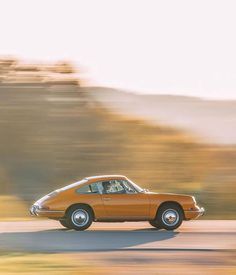 an orange car driving down the road in front of some hills and trees on a sunny day