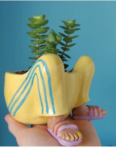 a hand holding a yellow ceramic planter with blue lines on the legs and feet