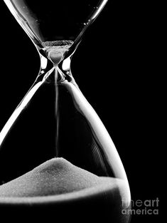 black and white photograph of an hourglass with sand running through the bottom, in front of a dark background