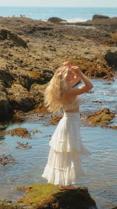 a woman in a white dress standing on rocks near the ocean with her hands to her head