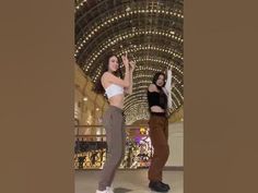 two women standing in front of a building with lights on the ceiling and one woman is holding her hand up