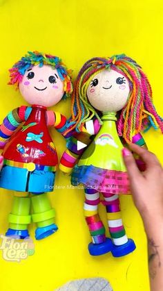 two colorful dolls sitting next to each other on a yellow surface with one being held up by someone's hand