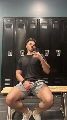 a man sitting on a bench in front of lockers taking a selfie with his cell phone