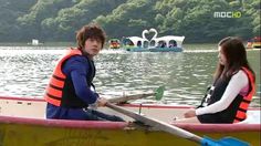 two people sitting in a boat on the water with swans floating behind them and one person wearing a life jacket