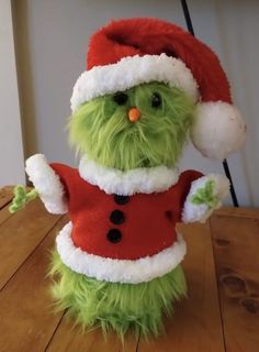 a green stuffed animal wearing a santa hat and red shirt on top of a wooden table