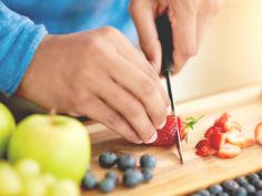 a person cutting strawberries with a knife on a cutting board surrounded by blueberries and apples