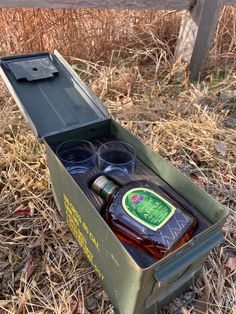 an open box with two glasses and a bottle in it sitting on the ground next to some dry grass