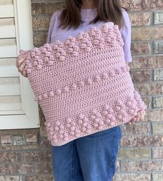 a woman holding a pink crocheted pillow in front of a brick wall