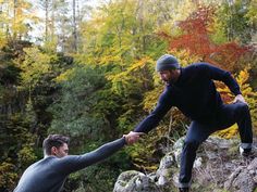 two men holding hands while standing on rocks in the woods