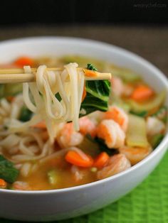 a white bowl filled with noodles and vegetables on top of a green place mat next to chopsticks