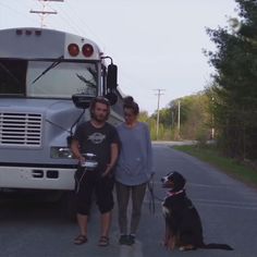 a man and woman standing next to a dog in front of a bus on the road