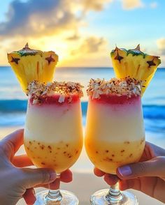 two people holding up glasses with drinks in them on the beach at sunset or sunrise