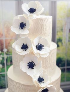 a three tiered cake with white and black flowers on the top, sitting in front of a window