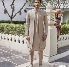 a man standing in front of a white building wearing a beige outfit and coat with an intricate pattern