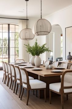 a dining room table with white chairs and vases on top of it, in front of large windows