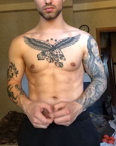 a man with tattoos on his chest standing in front of a bed and looking at the camera