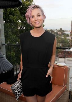 a woman with pink hair and black dress posing for the camera in front of a couch