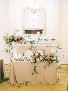 a table with flowers and candles on it in front of a fireplace decorated with greenery