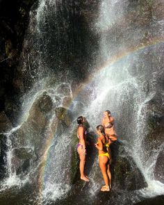 three women in bathing suits standing at the base of a waterfall with a rainbow behind them