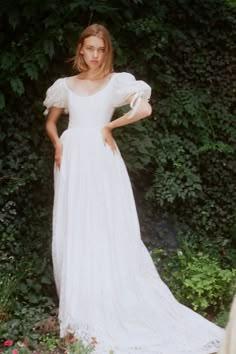 a woman wearing a white dress standing in front of some bushes