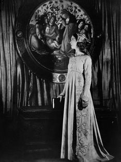 an old photo of a woman standing in front of a mirror with paintings on the wall behind her