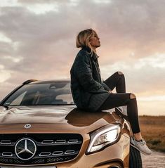 a woman sitting on the hood of a gold mercedes benz coupe parked in front of a field