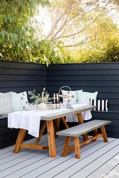 an outdoor table and bench with white linens on it, in front of a black wall