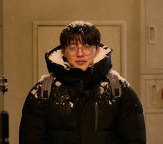 a man wearing glasses and a black jacket standing in front of a door with snow on it