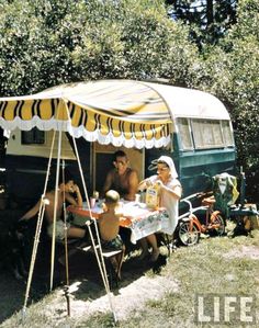 some people sitting under a yellow and white tent in the grass next to an rv