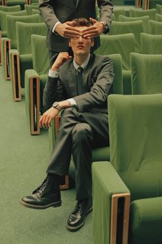 a man in a suit and tie sitting on a green chair with another man standing behind him