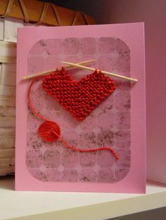 a pink card with a red heart hanging on a clothes line and knitting needles next to it