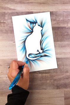 a person is drawing a cat with blue and white colors on the paper while holding a pen in their left hand