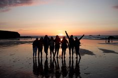 a group of people standing on top of a beach next to the ocean at sunset