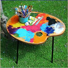 a colorful table with paint and brushes on it in the grass next to a tree