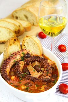 a bowl of soup with bread, tomatoes and olives next to a glass of wine