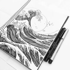 a spiral notebook with an ink drawing of a wave on it and two pens next to it