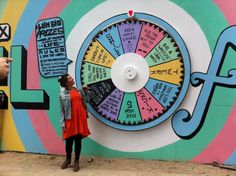 a woman standing next to a colorful wall with a wheel of fortune on it