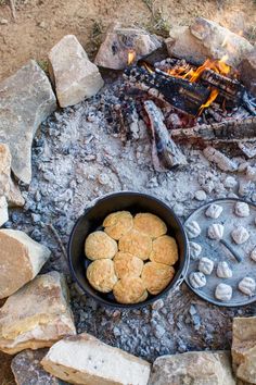 some biscuits are cooking over an open fire in the middle of rocks and gravel, with a campfire in the background